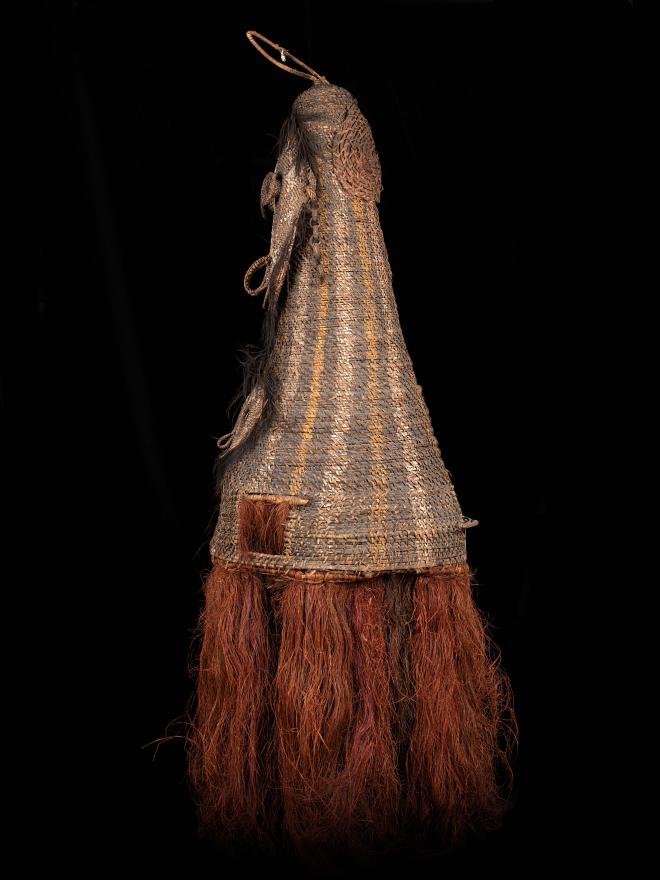 A double faced, tall woven dance mask from Kanganaman Village. The mask has two hand holes, a raffia skirt, and two holes in the lower face for the dancer to see out. Side View.