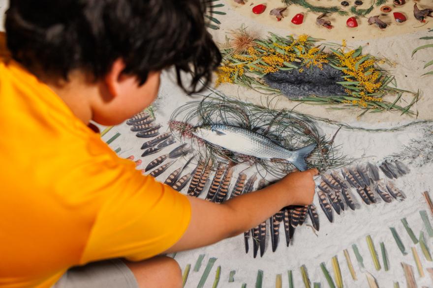 A young boy touches a large rug covered in natural materials like feathers, leaves and rocks