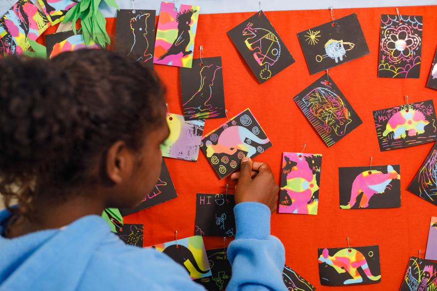 A young Aboriginal girl engaging with brightly coloured artworks featuring Australian animals pinned to a red wall.