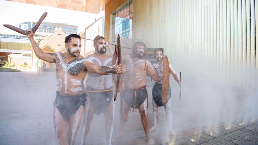 Four Aboriginal men in traditional paint and holding shields and Boomerangs, stand in a mist outside of the WA Museum