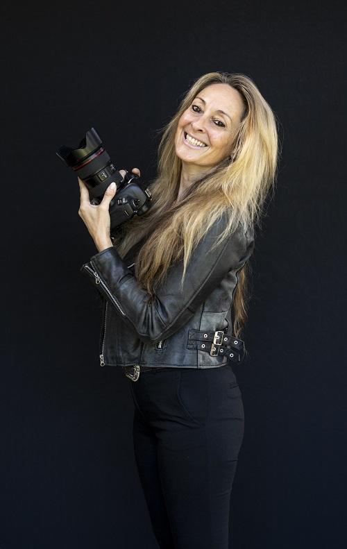 Photograph of Martine Perret, who has long blonde hair and wears a black leather jacket, posting in front of a black background with her camera in hand