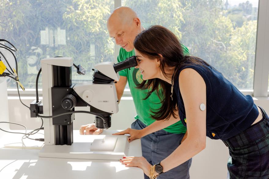 Chelsea, who wears a blue woolen vest, looks down the lens of a microscope as Nikolai watches on
