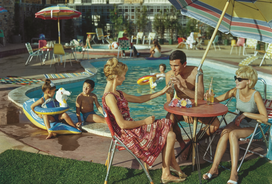 A vintage coloured photo of three people sitting under a pool umbrella at a small round table while their kids play in a motel pool behind them.