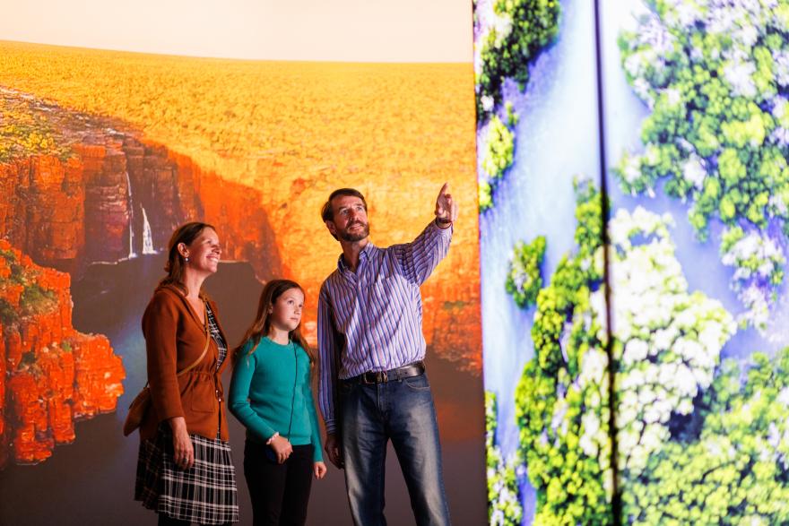 A family observes a large display featuring red cliffs and lush greenery over water.