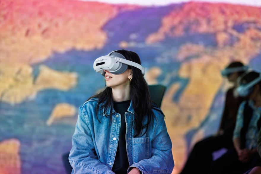 Person wearing a VR headset in front of a colourful landscape mural, with others in the background.