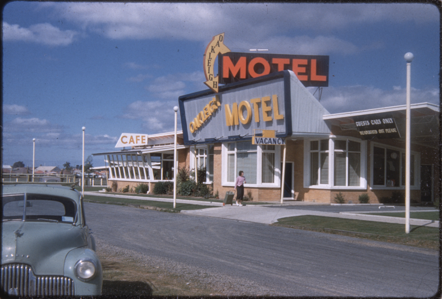 Vintage colourised photo of the Oakleigh Motel, taken from across the street. The motel is grey with large signage.