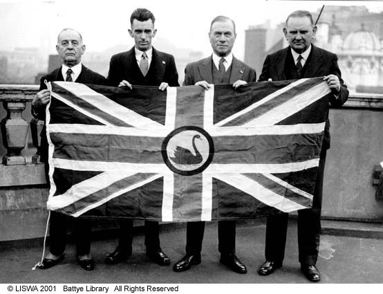 Four men holding a large flag displaying. The union jack stretches across the flag, with a black swan in a circle at the centre.
