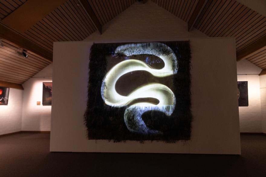 An illuminated abstract art piece on a wall with a textured dark border and a glowing yellow serpentine shape.