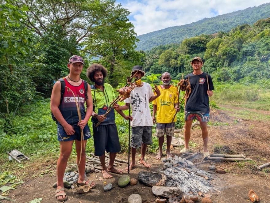 Five Indigenous young people standing around a small fire holding sticks with meat while surrounded by greenery