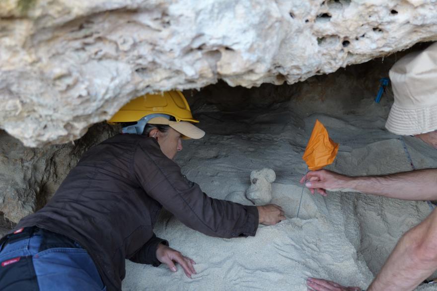 Person wearing a yellow hard hat reaches into a sandy cave area with exposed objects, while another hand holds an orange marker flag.