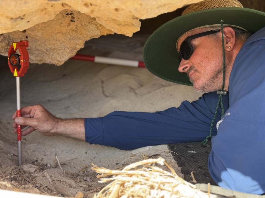 Person in a wide-brimmed hat using a measuring tool under a rocky overhang with sand and excavation equipment.
