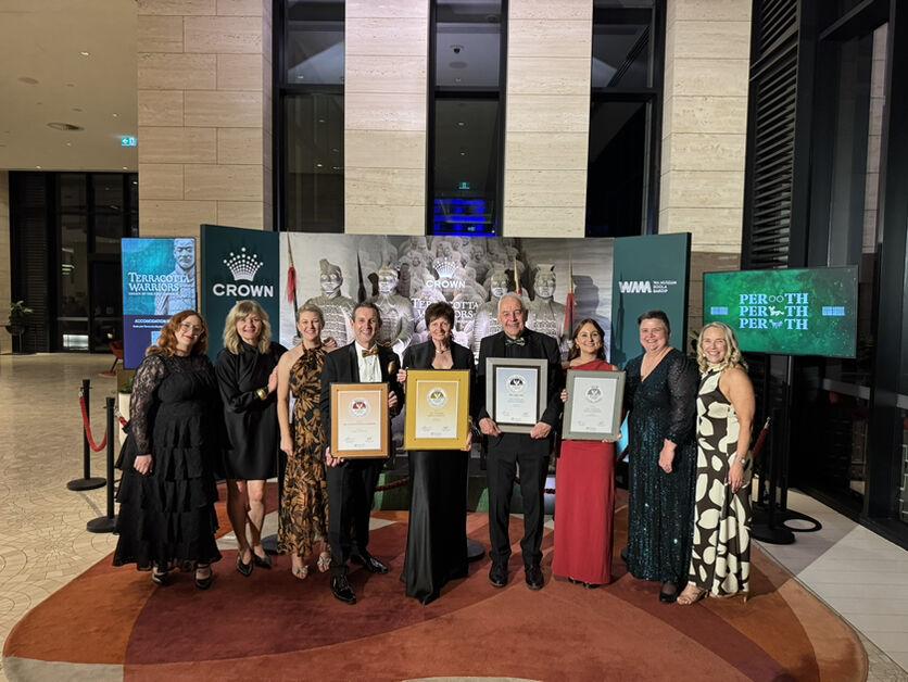 A happy group of 10 people from the WA Museum holding award certificates Infront of the Tourism award media wall after receiving an award