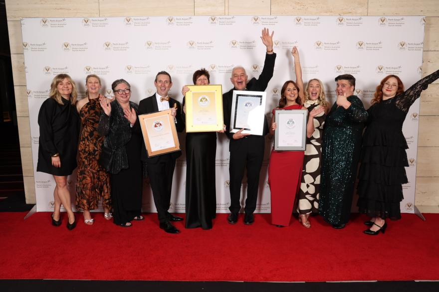 A happy group of 10 people from the WA Museum holding award certificates Infront of the Tourism award media wall after receiving an award