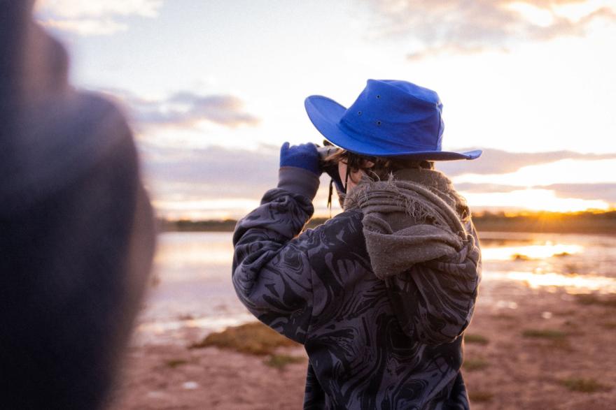 Person wearing a blue hat and patterned jacket looking through binoculars near a body of water at sunset.
