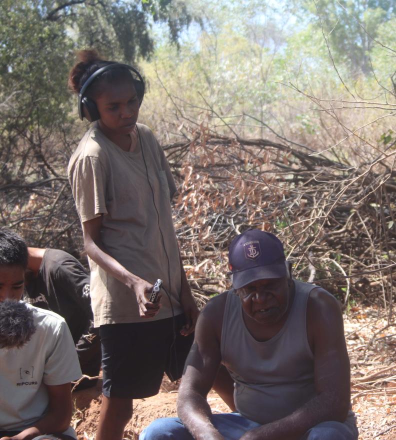 Group of people outdoors among dry branches and trees; one person wearing headphones and holding a small audio recorder.