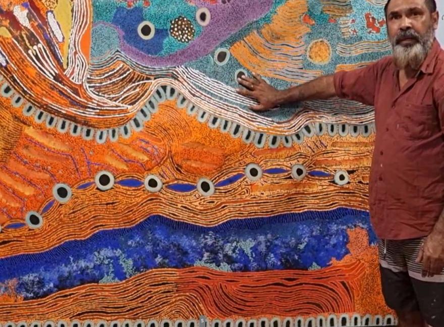Person standing beside a large, colourful Aboriginal artwork featuring intricate patterns in orange, blue, purple, and teal.