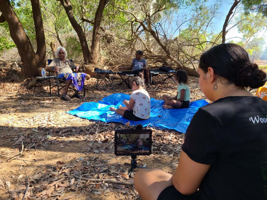 Group of people sitting on a blue tarp in a wooded area, with musical keyboards on stands and a camera recording the scene.