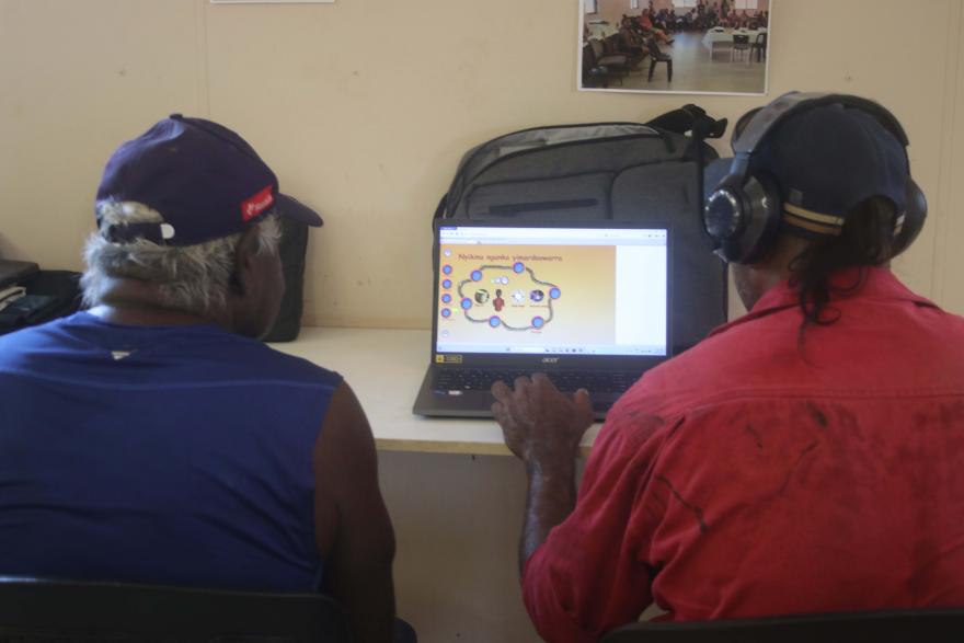 Two people sitting at a desk looking at a laptop screen displaying an educational game with text and colourful icons