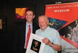 Two people standing indoors near a display banner that reads “Western Australian Museum.” One person is holding a framed image of a brightly colored sea creature. Behind them are dark walls with vibrant artwork mounted.