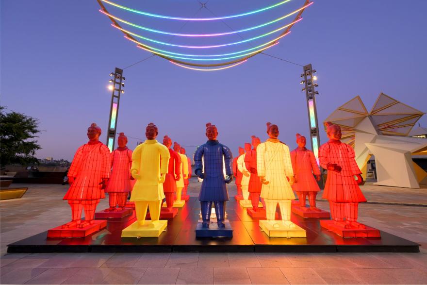 Colourful illuminated lanterns resembling Terracotta Warriors arranged in rows, with glowing neon arcs overhead at dusk.