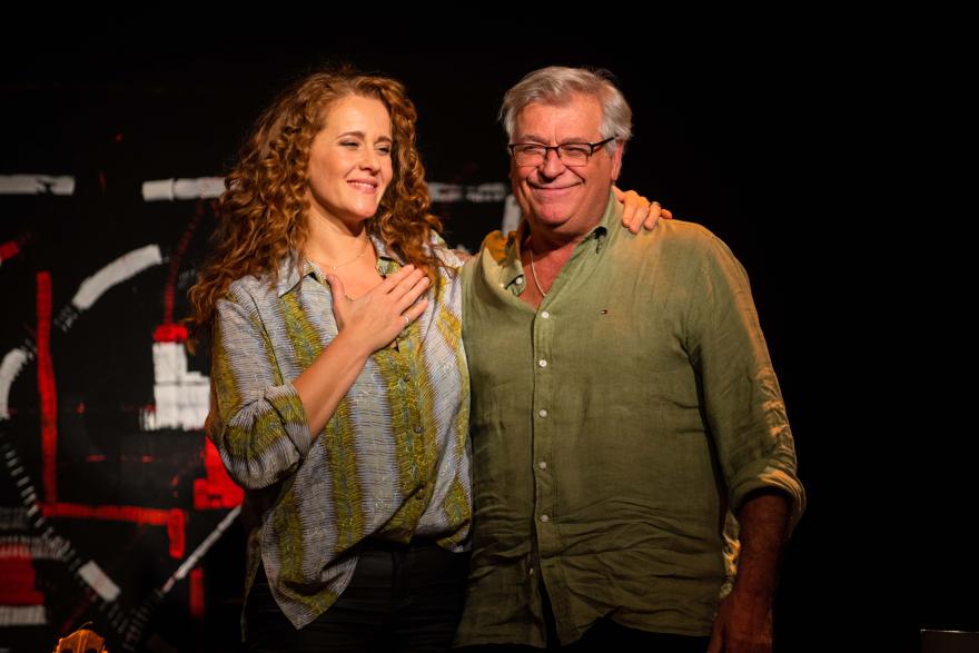 Two people standing together onstage, one with a hand on their chest and the other with an arm around them.