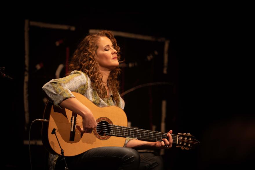 A musician seated onstage playing an acoustic guitar under warm stage lighting.