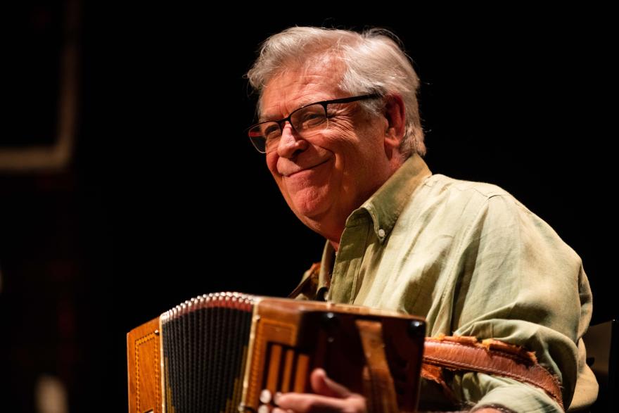 A musician seated onstage playing an accordion, lit by a spotlight.