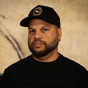 a headshot style photo where an adult person looking at the camera. They wear a black hat and a black tshirt