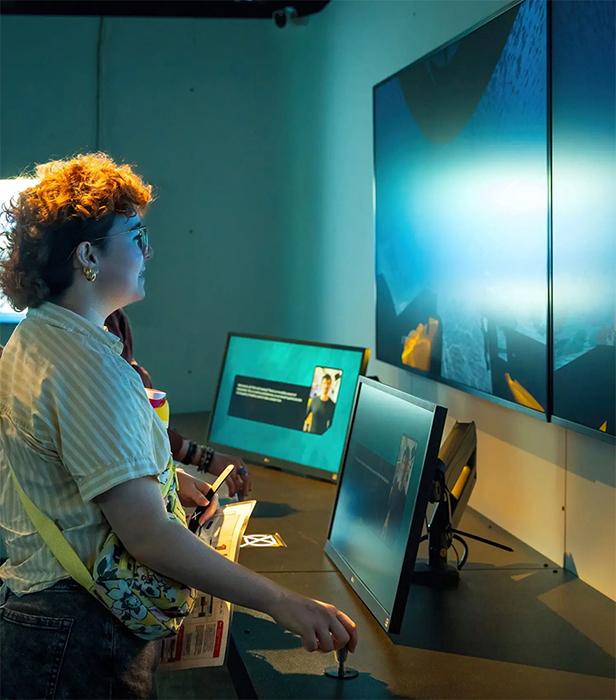 A person interacts with a digital exhibit, using joystick-style controls while watching large screens that display underwater scenes.