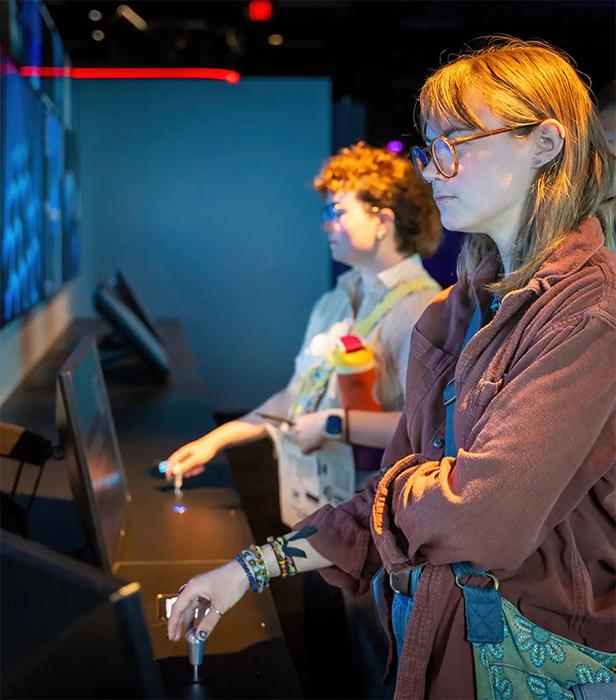  People standing at an interactive exhibit, using joystick-style controls while watching large screens displaying underwater visuals.