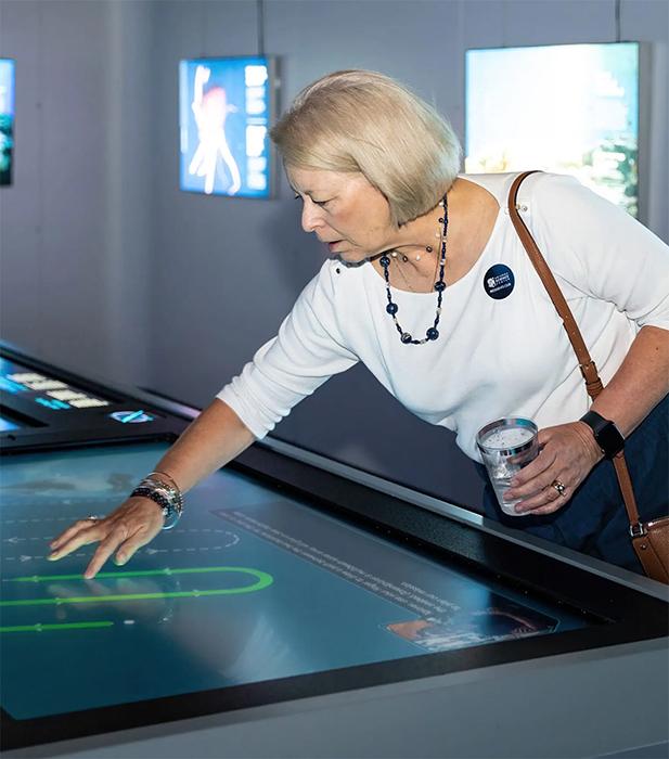 A person with light hair interacts with a large touchscreen display in a dimly lit exhibition space, using one hand to navigate the on‑screen content while holding a drink in the other.