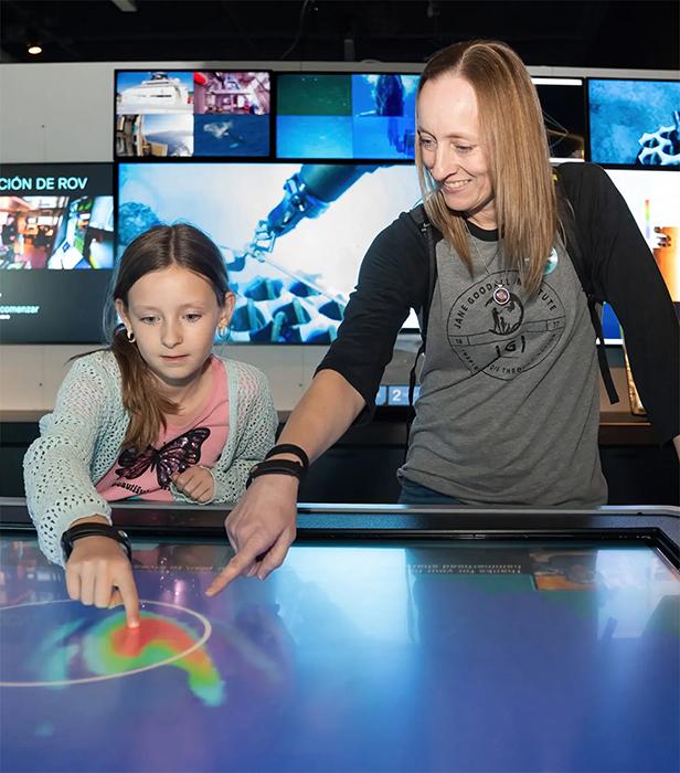 Two people interact with a large touchscreen display, pointing at colourful graphics while multiple screens in the background show underwater and ROV footage.