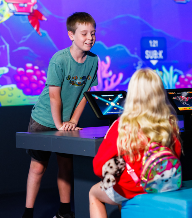 Two children interact at a touchscreen exhibit with a colourful underwater-themed display in the background.
