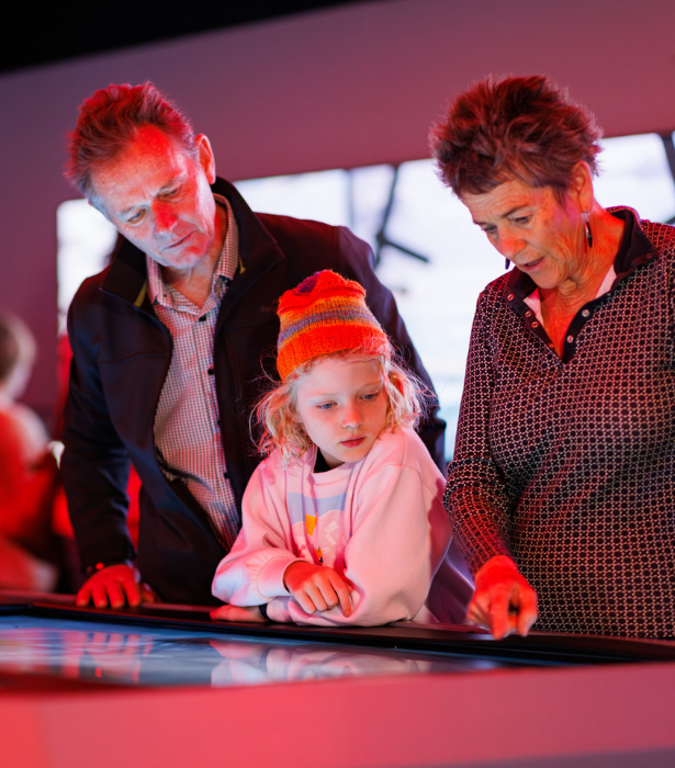 An adult and a child use an interactive touchscreen table together in a museum-style exhibit with red lighting.