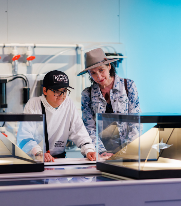 Two people engage with a hands-on display at an interactive museum exhibit, with glass cases and lab-style equipment nearby.