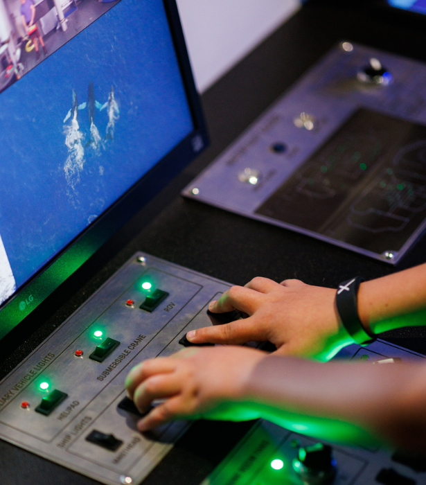 Hands operate a lit control panel at an interactive exhibit, with a screen showing marine animals above the controls.