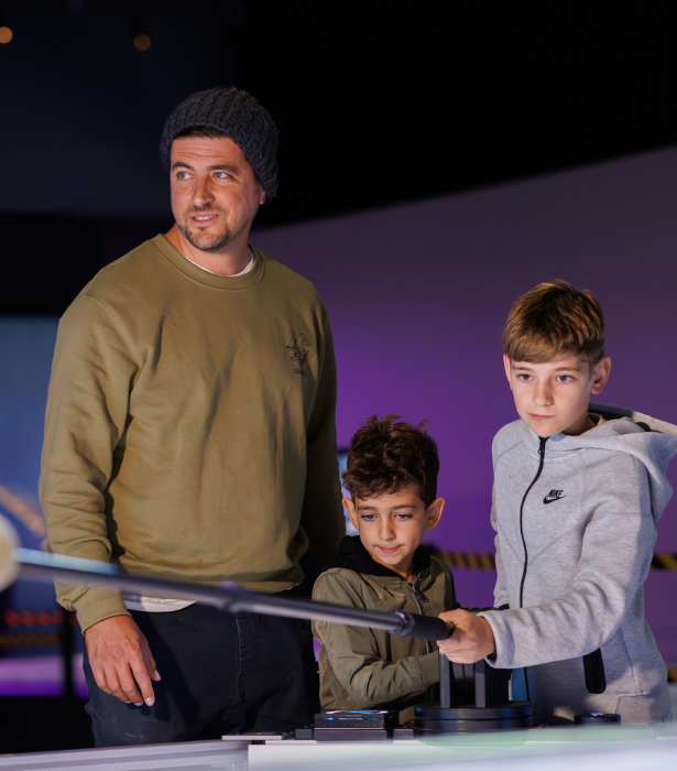 An adult and two children use a hands-on exhibit, with one child holding a long lever over an interactive display.