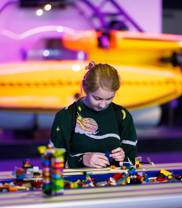A child builds with LEGO pieces at a long activity table, with a brightly lit yellow boat display in the background.