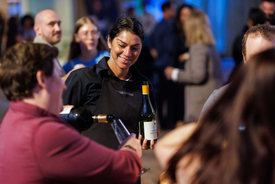 A person pours wine into a glass at a crowded indoor event, with people mingling in the background.