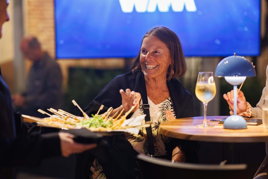 A person sits at a small round table at an indoor event, gesturing while a server offers a tray of small snacks, with drinks and a crowd in the background.