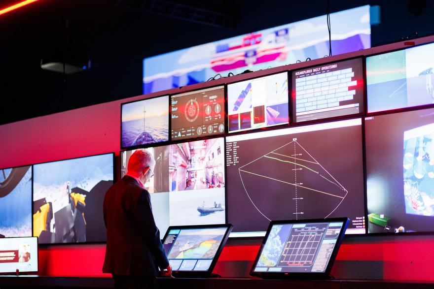 A person stands at a control station facing multiple large screens showing ocean data, maps, and video feeds in a dimly lit room.