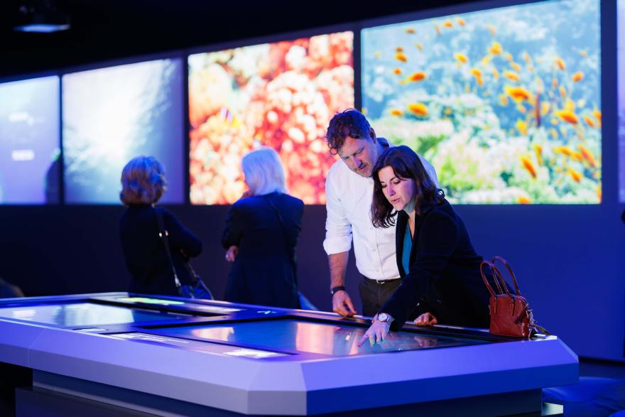Visitors interact with a large touchscreen table displaying digital content, with illuminated ocean imagery panels on the wall behind them.