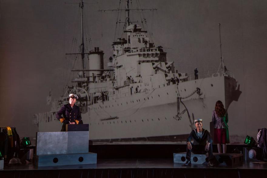 a person on stage with a sailors uniform on a podium with a projected image of a ship in the background