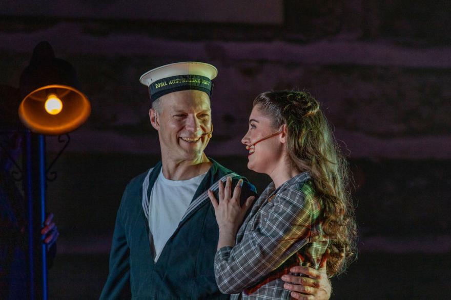 two adults looking at eachother holding eachother in a half hug and smiling at eachother. One person wears a sailors outfit and another person is in a checkered blazer