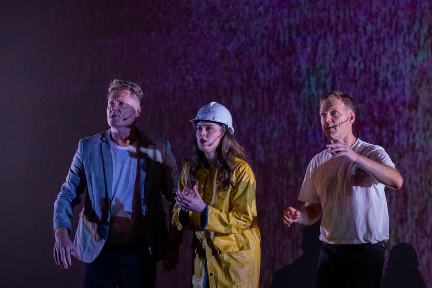 three people staring out into with concerned looks on their faces. One person wears a t shirt and grey blazer, another person wears a yellow rainjacket and hardhat and the last person wears a white tshirt