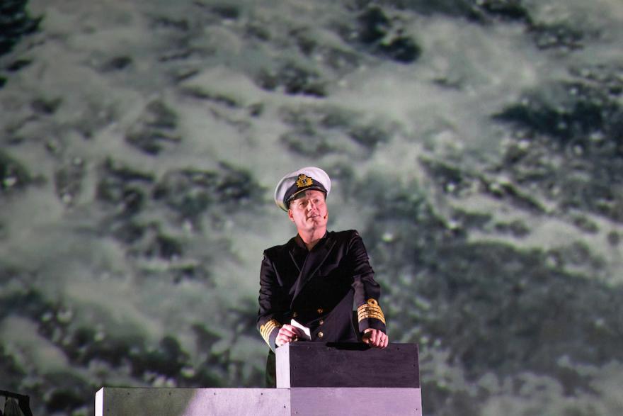 a person in a sailing uniform stands looking out with an ocean wave projection in the background