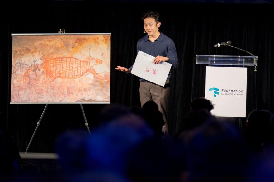 A man on stage in front of a screen displaying Aboriginal rock paintings