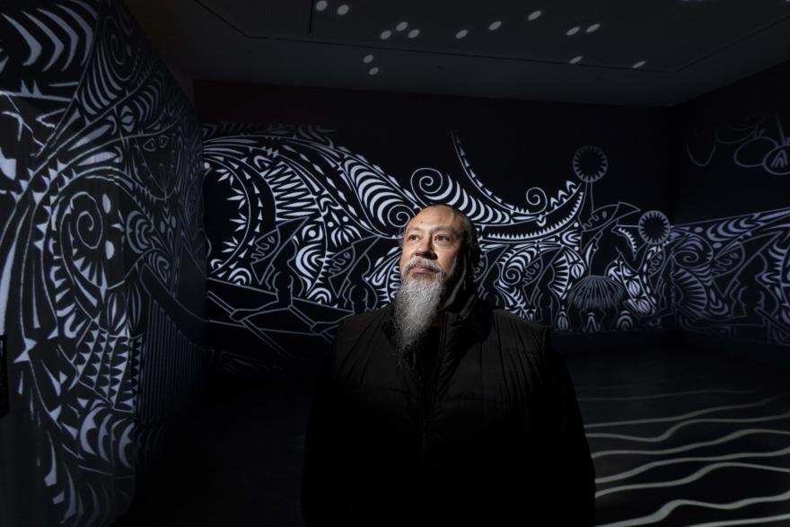 A person stands in a dark gallery space illuminated by a black-and-white projected artwork featuring bold, swirling patterns and abstract motifs covering the walls and floor.