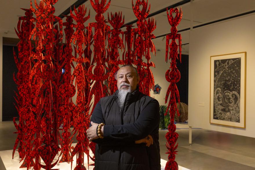 A person stands with folded arms in an art gallery beside a large red hanging installation made of intricate patterned cut-outs, with additional framed and sculptural artworks displayed in the background.