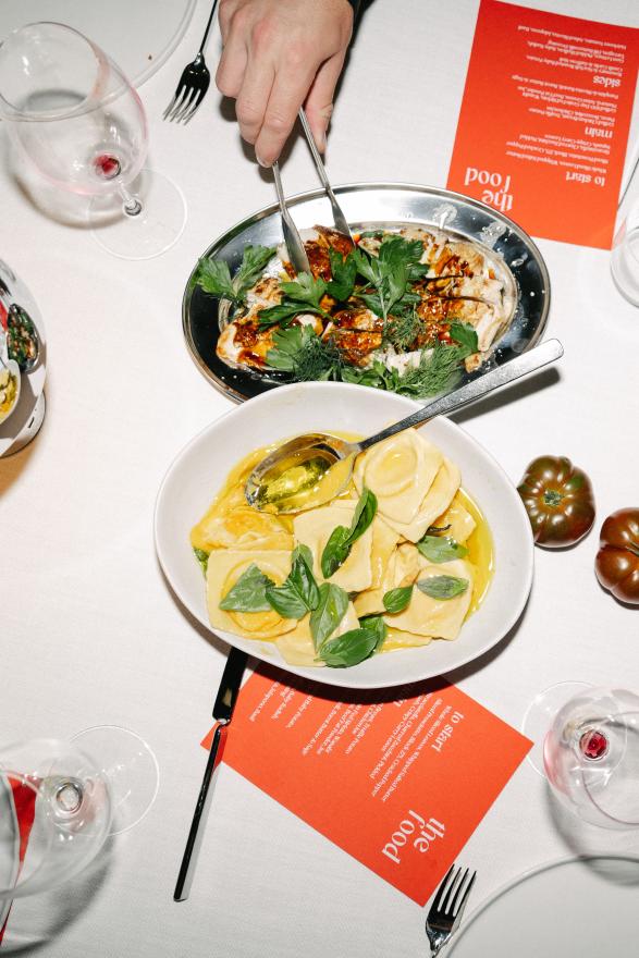 Overhead view of a restaurant table with a bowl of ravioli in butter and herbs, a plated dish garnished with greens, a hand using serving utensils, wine glasses, and red menus on a white tablecloth.